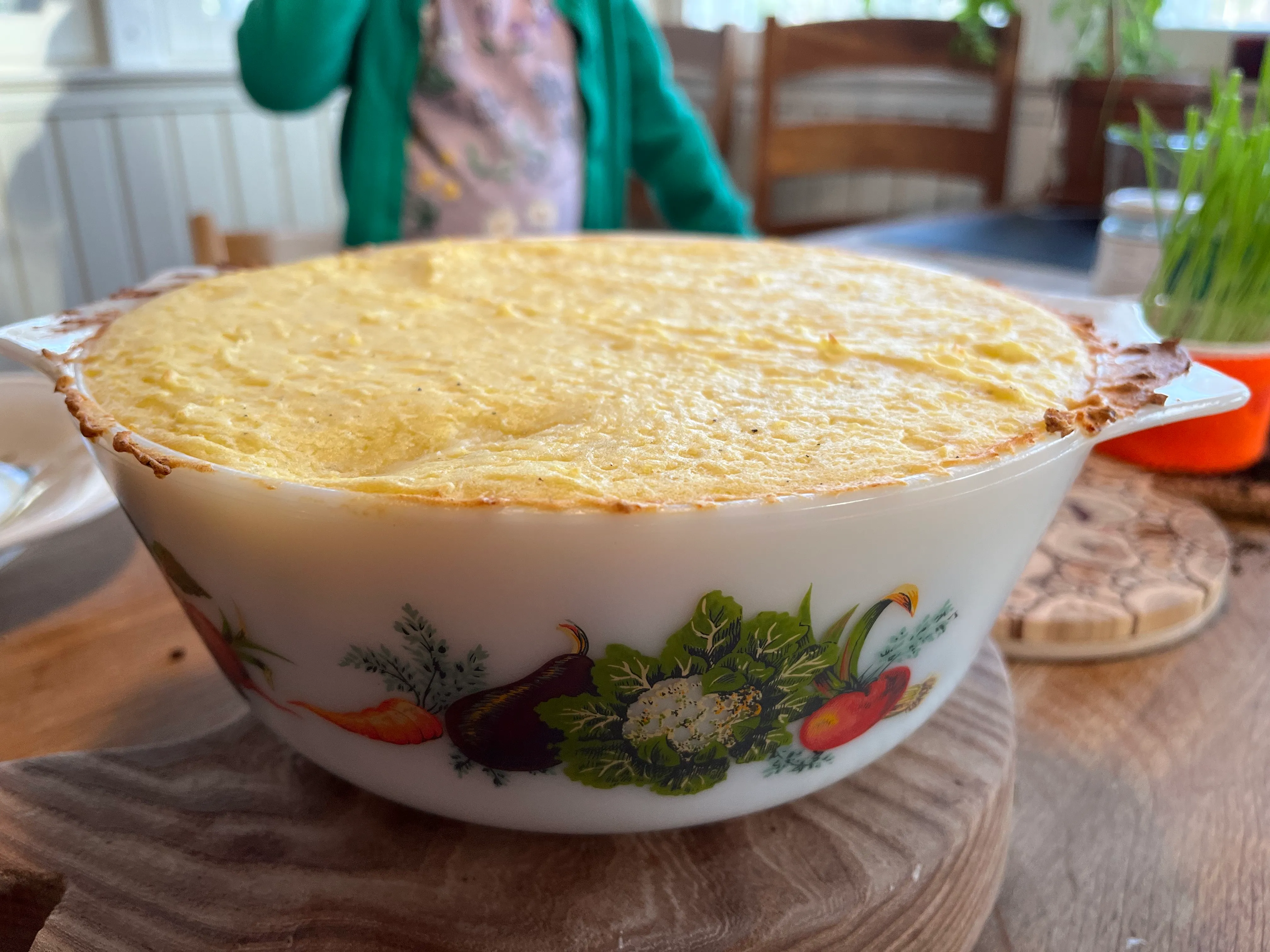 A golden-topped baked casserole in a vintage vegetable-print dish resting on a wooden board