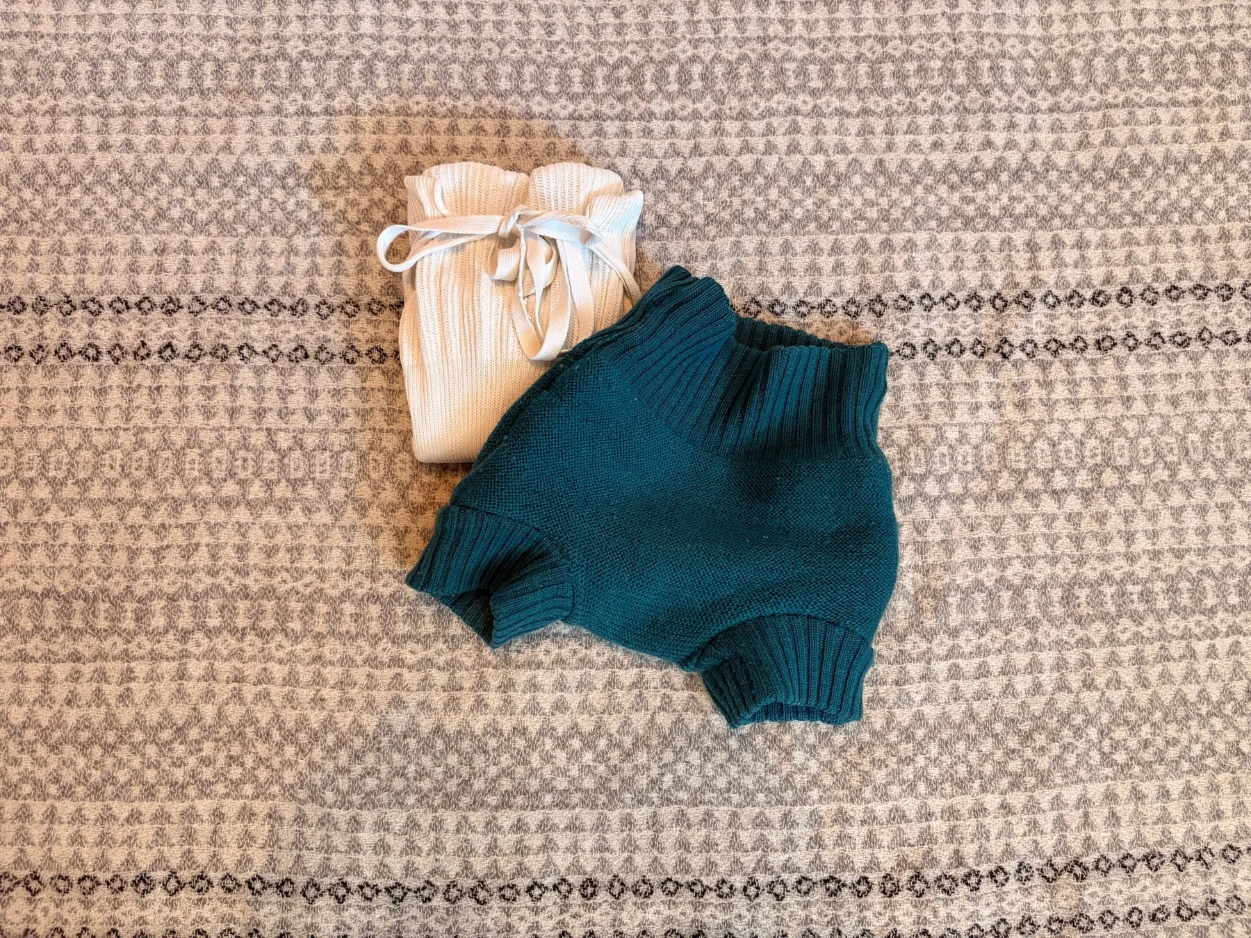A folded cotton tie nappy beside a teal knitted wool cover on a patterned rug